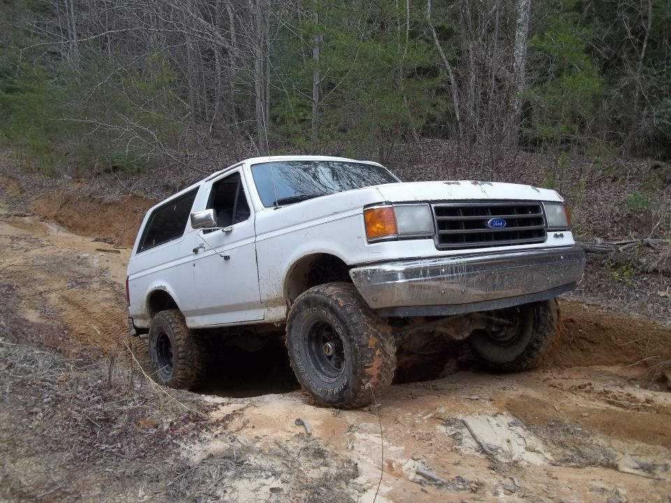 White Ford Bronco with lifted suspension driving on a dirt trail through woods