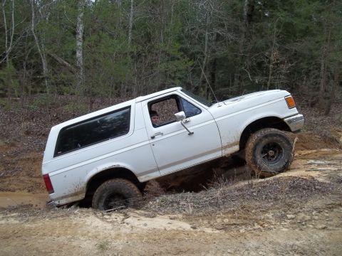 1990 Ford Bronco - A white 1990 Ford Bronco is shown driving off-road through a muddy terrain.