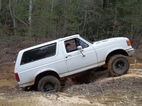 1990 Ford Bronco - A white 1990 Ford Bronco navigates a challenging off-road course, with its tires digging into the muddy terrain.