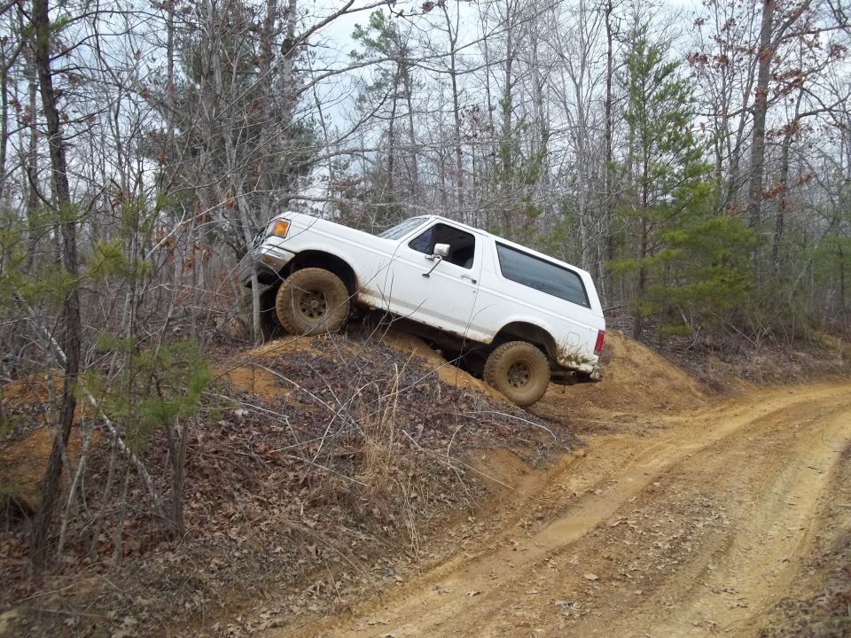 White 1992 Ford Bronco climbing a dirt hill off-road, front-left tire elevated