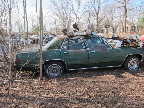 1991 Ford LTD Crown Victoria - This green sedan, likely from the 1980s, is in a state of disrepair at a junkyard, with parts stacked on its roof.