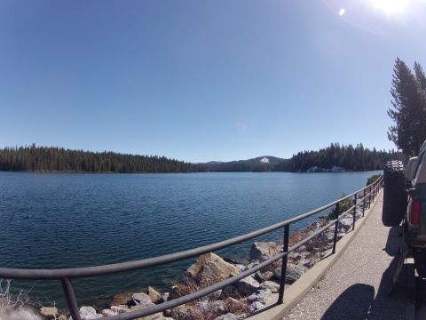 1989 Ford Bronco - A vehicle is parked on a paved path next to a railing overlooking a calm lake surrounded by pine trees under a clear blue sky.
