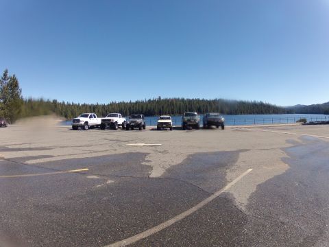 1989 Ford Bronco - Several SUVs and trucks are parked in a lot overlooking a lake, with a dense forest in the background under a clear blue sky.
