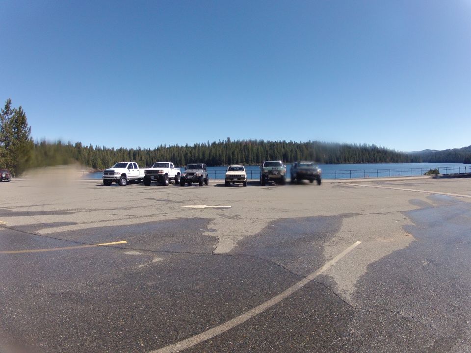 Group of SUVs and trucks parked by a lake with a forest and blue sky