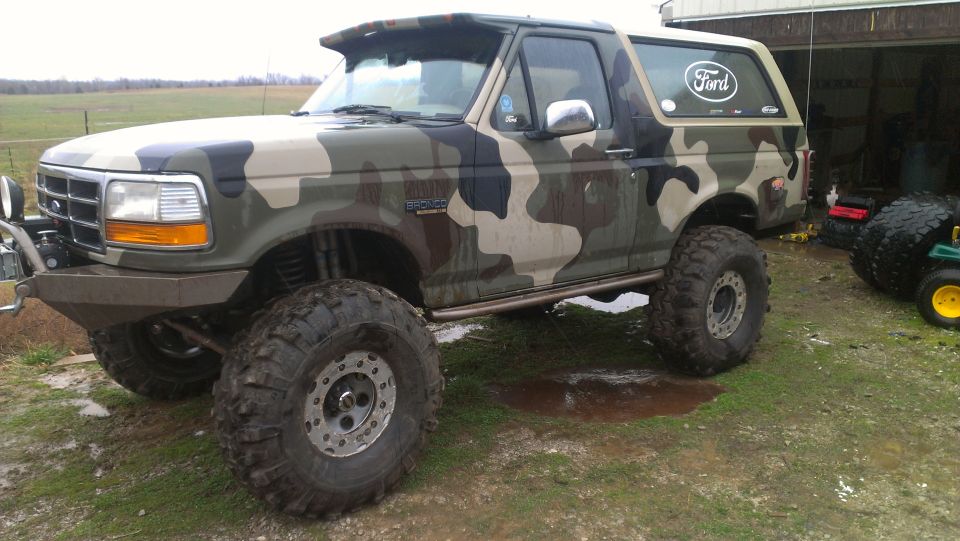 1995 Ford Bronco with camouflage wrap and large off-road tires parked on wet ground