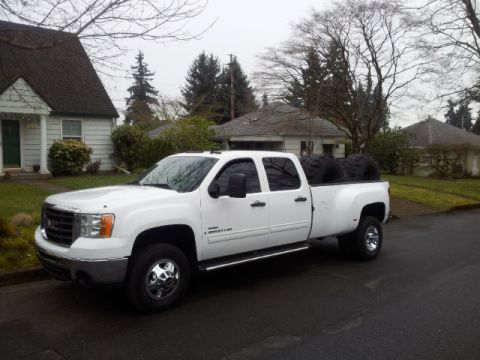 2008 GMC Sierra 3500 - A white GMC Sierra 3500 dually pickup truck is parked on a street in front of houses, with spare tires loaded in the bed.