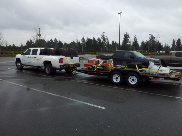 White dually pickup truck towing a flatbed trailer carrying a black pickup truck and cargo