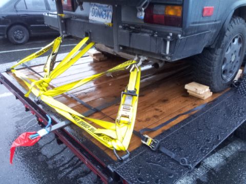 2008 GMC Sierra 3500 - A dark van is secured with yellow straps to a flatbed trailer, with a black car visible in the background.