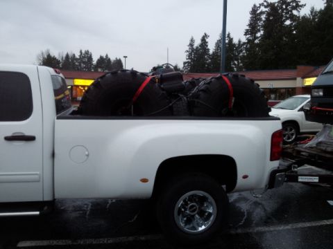 2008 GMC Sierra 3500 - Pickup truck carrying oversized tires, with other vehicles and buildings in the background.