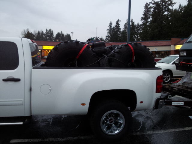 White pickup truck with large tires strapped in the bed. Another vehicle is visible in the background