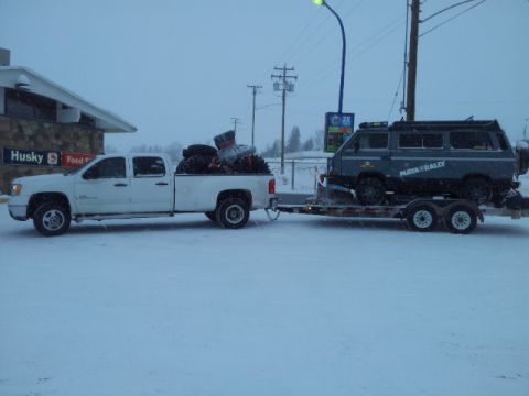 2008 GMC Sierra 3500 - A white pickup truck tows a trailer carrying a gray van through falling snow.