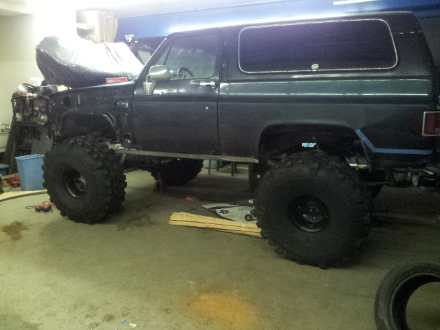 Dark-colored SUV with large off-road tires, lifted suspension, and hood removed, sitting in a garage