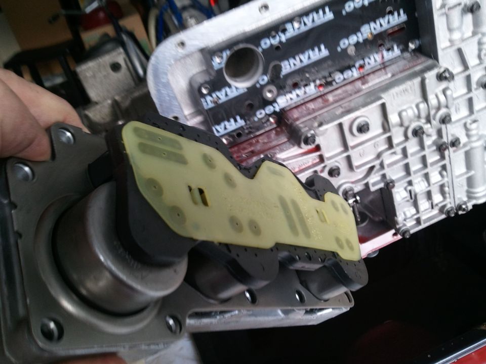 Transmission valve body with "TRANSTEC" printed on it. A hand is holding a part of it