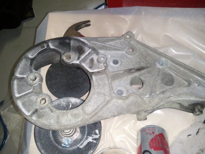 Close-up of a metal automotive part, possibly a bracket, with a hammer and grinding discs nearby. A Supermotors.net logo is visible