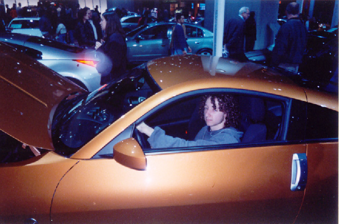 Person seated in orange concept coupe at an indoor auto show, crowd and cars behind