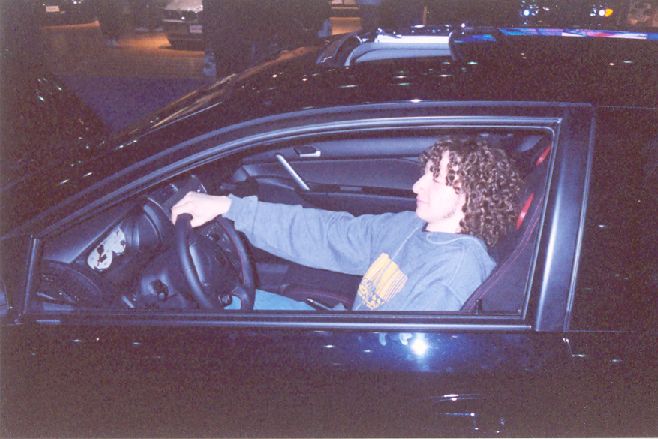 Person with curly hair seated at the wheel of a dark car, viewed through the open driver's window at an indoor show