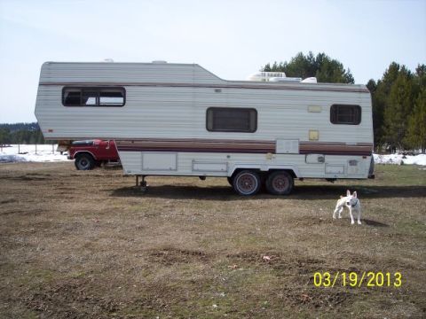 1973 Ford F-250 - A fifth wheel trailer and a red pickup truck are parked in a field on a sunny day, with a dog standing nearby.