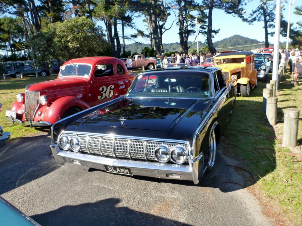 Front view of a black 1963 Lincoln Continental with a custom grille and license plate that says SLAMM. Red car with number 36 is to the left
