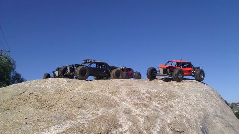 1996 Ford Bronco - Multiple RC off-road vehicles are positioned on a large rock formation against a bright blue sky.