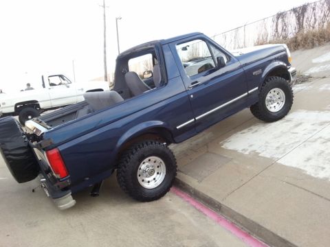 1995 Ford Bronco - Dark blue 1995 Ford Bronco with its top removed is parked on a curb, with a white pickup truck behind it.