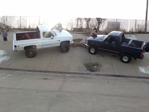 1995 Ford Bronco - Two lifted trucks, a 1995 Ford Bronco and a white pickup, are parked on a paved surface with people sitting on their hoods.