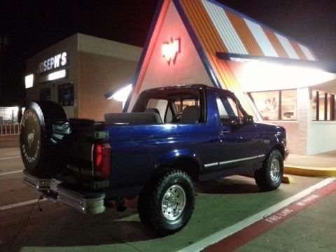 1995 Ford Bronco - Blue 1995 Ford Bronco with a spare tire on the rear is parked in front of a building at night.