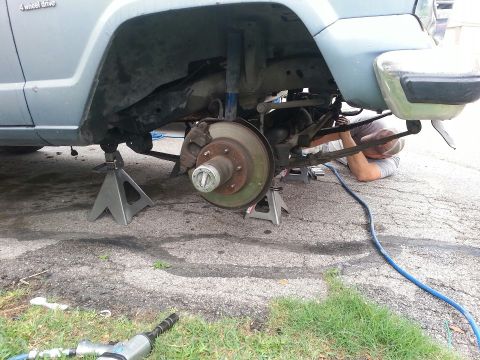 1982 Jeep Wagoneer - Mechanic works on the suspension of a light blue Jeep, which is raised on jack stands.