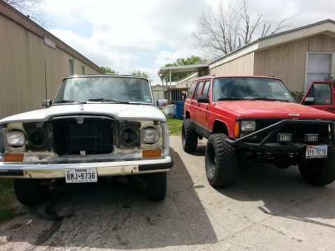 1982 Jeep Wagoneer - Two Jeep SUVs are parked side-by-side in a driveway with buildings in the background.