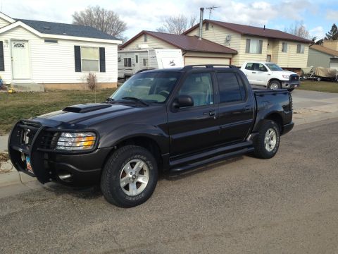 2005 Ford Sport Trac - A dark 2005 Ford Sport Trac is parked on a street in front of houses, with a white pickup truck in the background.