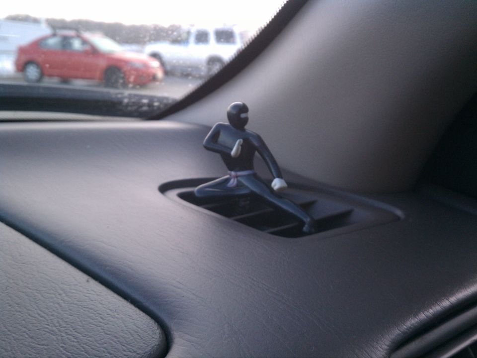 Dashboard vent with a ninja figurine, red car and white SUV visible through windshield