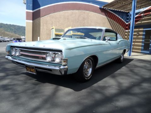 1973 Ford Ranchero - light blue Ford Torino GT fastback with quad headlights and styled wheels parked on pavement in front of a striped building.