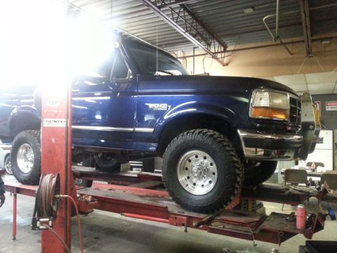 1995 Ford Bronco - A blue Ford Bronco is raised on a hydraulic lift inside a workshop, showing its undercarriage and suspension.