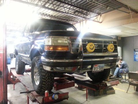 1995 Ford Bronco - A black 1995 Ford Bronco is raised on a lift in a garage, showing off its front grille with decorative smiley face lights.