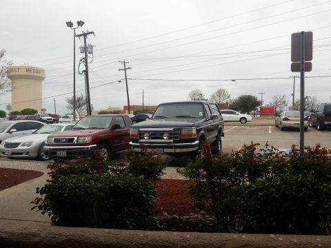 1995 Ford Bronco - A dark blue 1995 Ford Bronco is parked in a lot next to a red pickup truck.