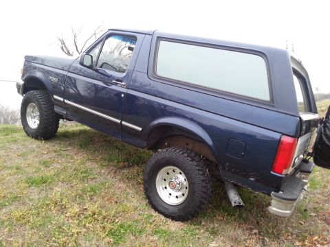 1995 Ford Bronco - This lifted 1995 Ford Bronco is parked on a grassy slope, showing its off-road tires and custom wheels.
