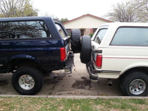 1995 Ford Bronco - Rear view of two Ford Bronco SUVs, one dark blue and one white, parked closely together.