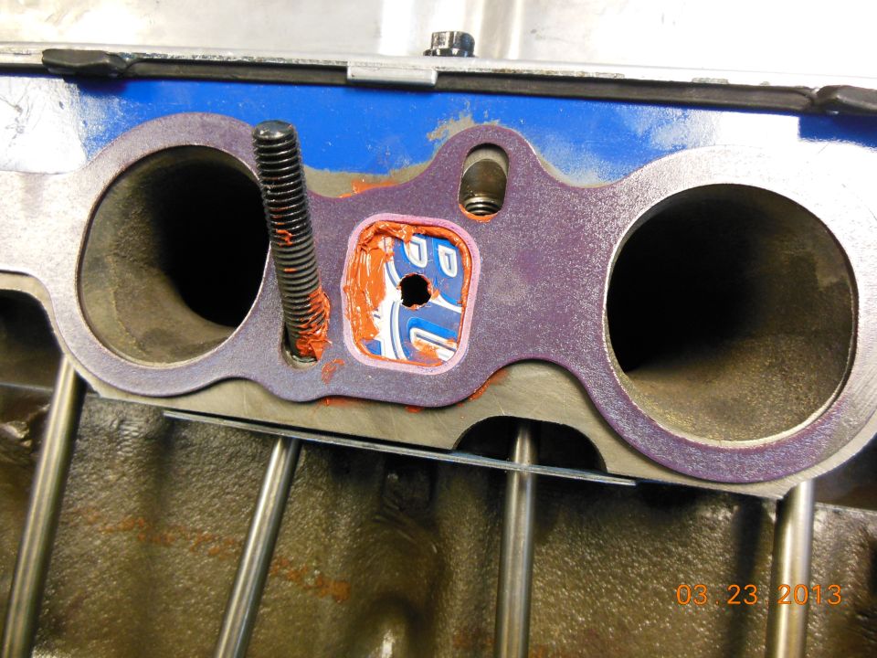 Engine manifold gasket with orange sealant and a bolt. Visible text reads 'SUPERMOTORS.NET'