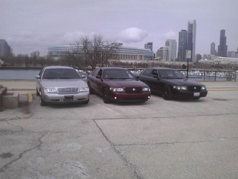 2003 Ford Crown Victoria - Three sedans are parked in a lot near a body of water with a city skyline in the distance.