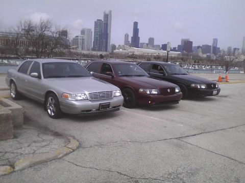 2003 Ford Crown Victoria - Three Ford Crown Victorias are parked in a lot with a city skyline visible behind them.