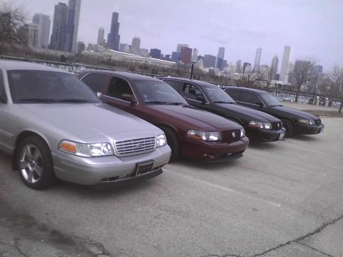 2003 Ford Crown Victoria - A line of Ford Crown Victorias, including a silver model in the foreground, are parked with a city skyline in the background.
