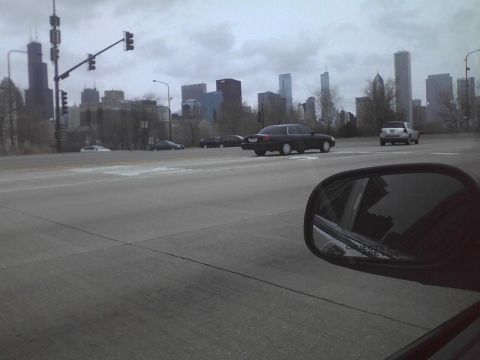 2003 Ford Crown Victoria - A dark sedan drives along a road with the Chicago skyline visible under a cloudy sky.