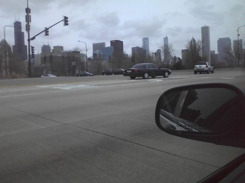 Dark sedan driving on a road with a city skyline in the background. Another vehicle is ahead