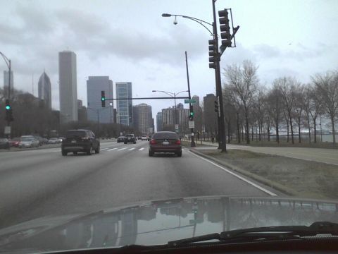 2003 Ford Crown Victoria - A dark sedan travels down a multi-lane road with a city skyline visible under an overcast sky.