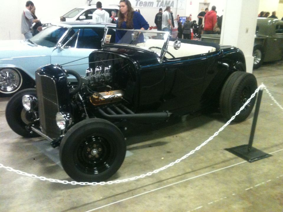 black hot rod roadster with exposed engine and wide rear tires, parked indoors with a light blue car and other people nearby