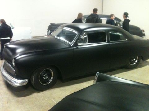2004 Ford Crown Victoria - A matte black hot rod coupe is displayed indoors, with other vehicles and people in the background.
