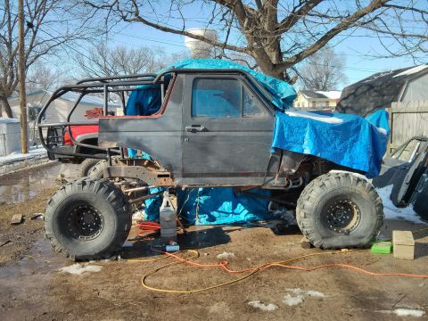 1986 Ford Bronco - This heavily modified SUV is in a state of repair or customization, with parts covered by blue tarps.