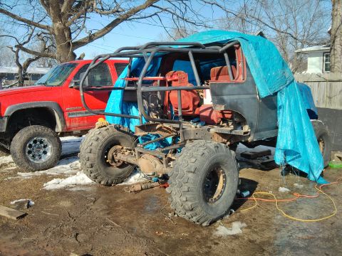 1986 Ford Bronco - Modified off-road vehicle chassis with a roll cage and blue tarp is parked next to a red pickup truck.
