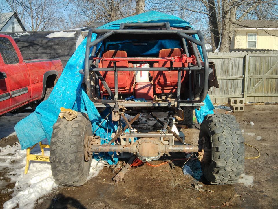 Off-road vehicle rear view with roll cage, red seats, and large tires. A red pickup truck is partially visible