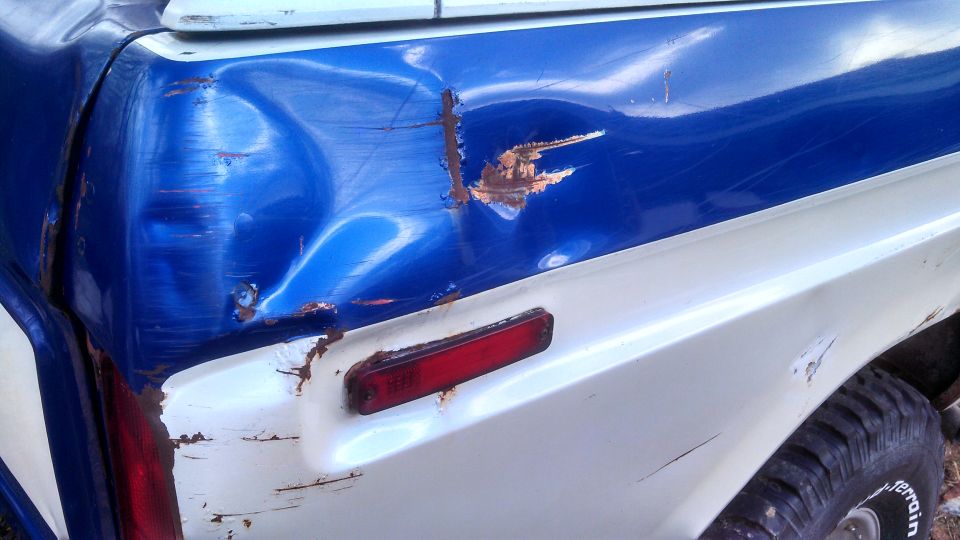 Close-up of a dented blue and white truck rear quarter panel with rust and a red taillight