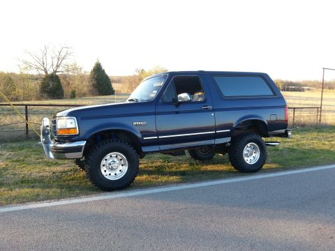 1995 Ford Bronco - A lifted blue Ford Bronco is parked on the shoulder of a road with a rural landscape in the background.